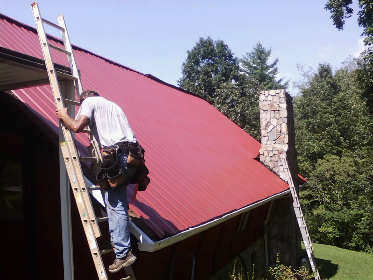 Roofer on a ladder beside a red metal roof and stone chimney — Soffit & Fascia Repair by Roofing Brothers in New Springfield